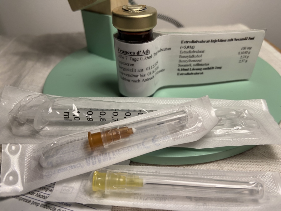 A close-up of a small vial of Estradiol valerate with a packaged syringe and two needles in front.